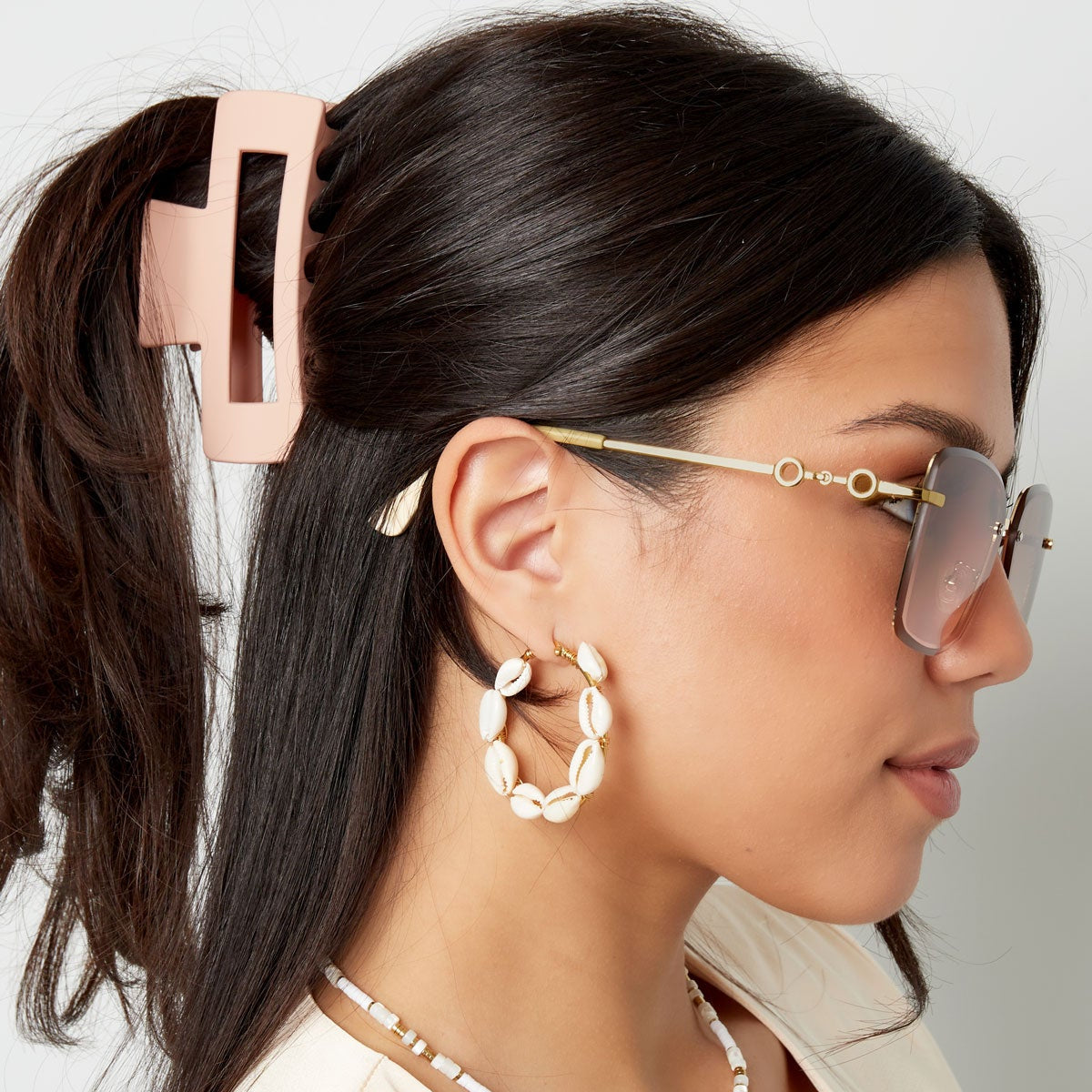 Woman wearing sunglasses, hoop earrings, and a necklace with a neutral background